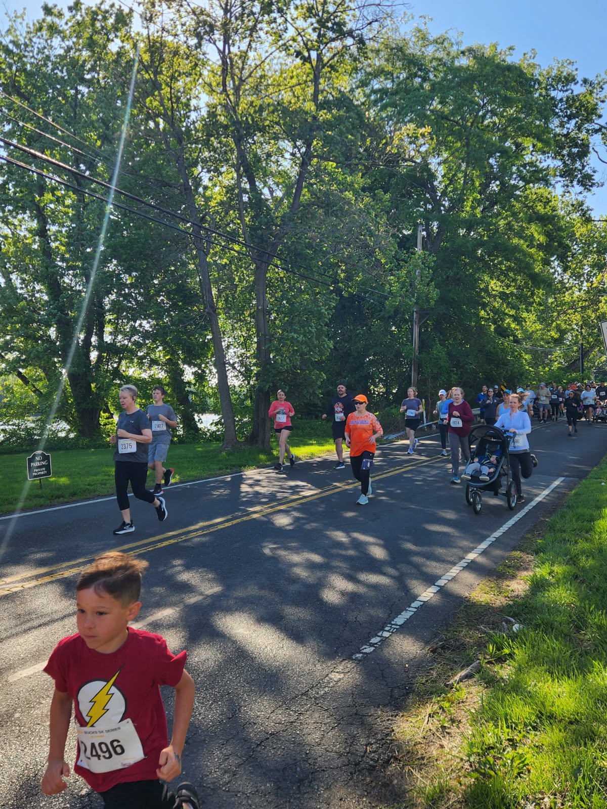 A young runner in a red shirt sprinting ahead of the pack along the tree-lined Yardley 5K course near the Delaware Canal
